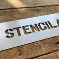 A rectangular white stencil sheet with the word 'STENCILAND' cut out, placed diagonally on a rustic wooden surface.