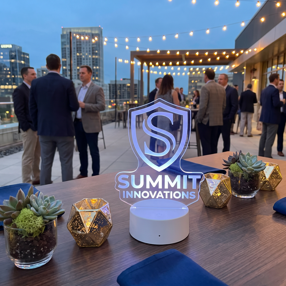 A rooftop event scene at dusk with several people mingling in the background, a wooden table in the foreground, and a glowing acrylic centerpiece that reads “SUMMIT INNOVATIONS.” Decorative gold geometric vases, small succulent plants in glass cups, and blue napkins are arranged on the table.