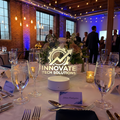 A formal event table setup in a brick-walled banquet hall, with clear wine glasses, silverware, napkins, and a central logo overlay reading 'Innovate Tech Solutions'. Guests are visible mingling in the background under warm string lights and blue accent lighting.